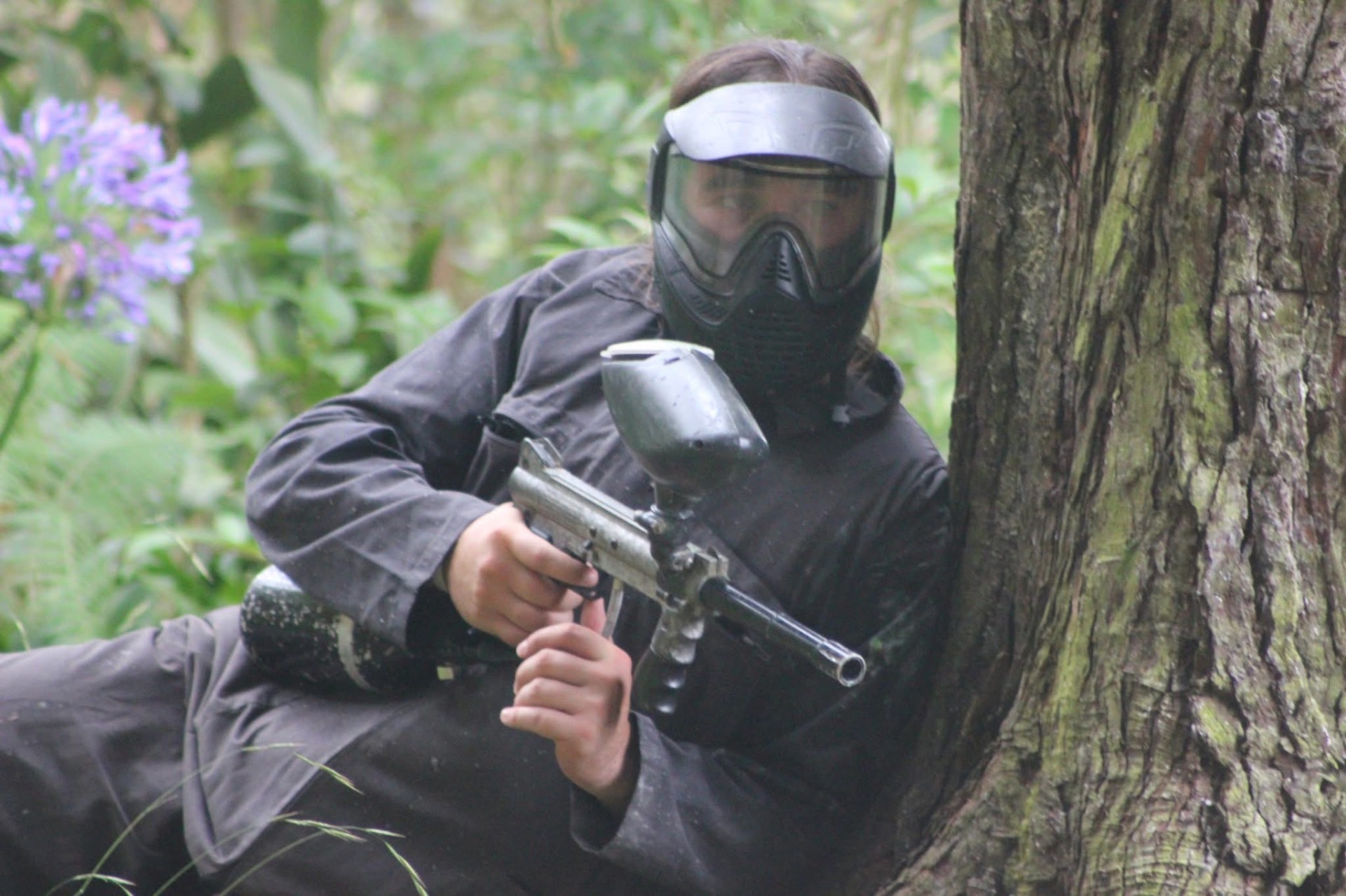 Jogador de paintball a apontar o marcador protegido por uma árvore